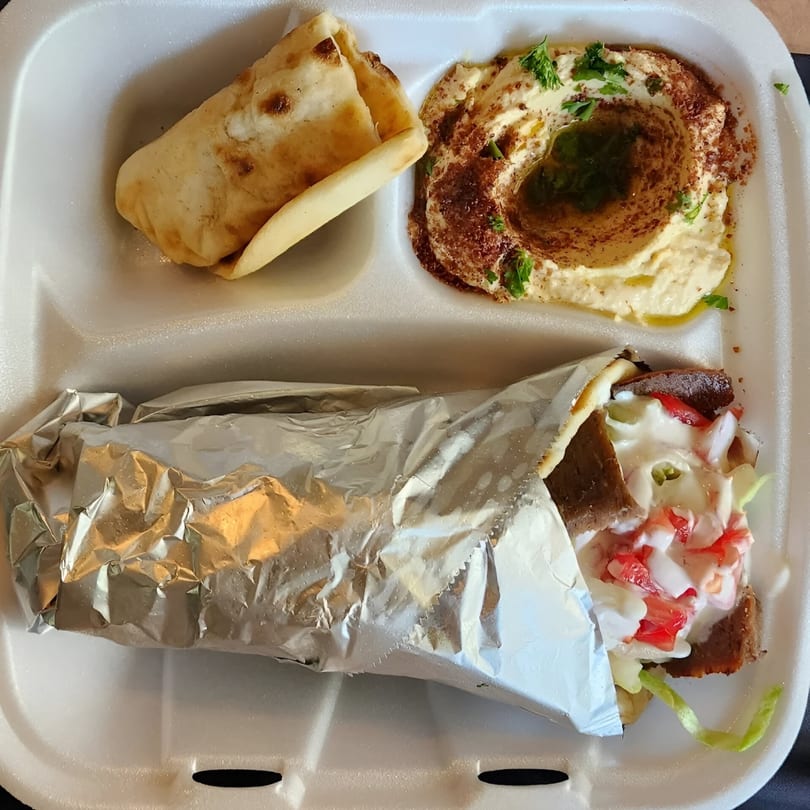 A Taza Grill Mediterranean meal with a gyro, hummus, and pita bread in a white takeout container.